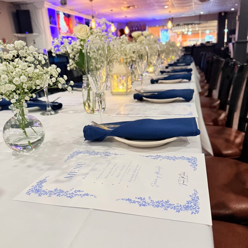 A long dining table set with white tablecloths, blue napkins, menus, and floral centerpieces at Franco Martini.