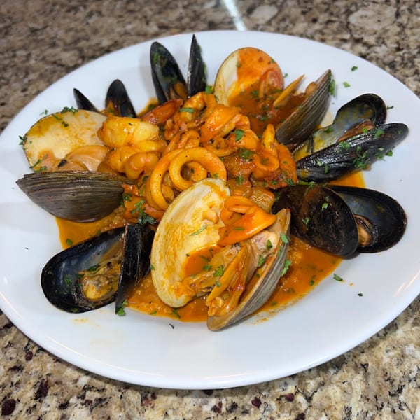 A white plate holds an Italian seafood dish with mussels, clams, and calamari in a rich red sauce.