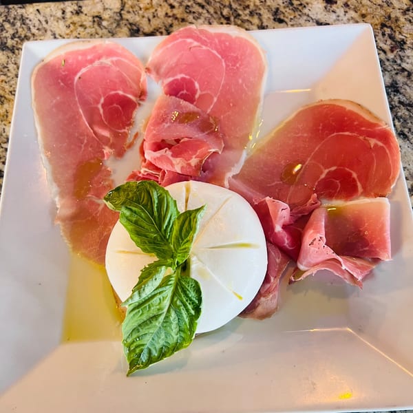 Italian prosciutto and burrata cheese with fresh basil and olive oil on a white plate.